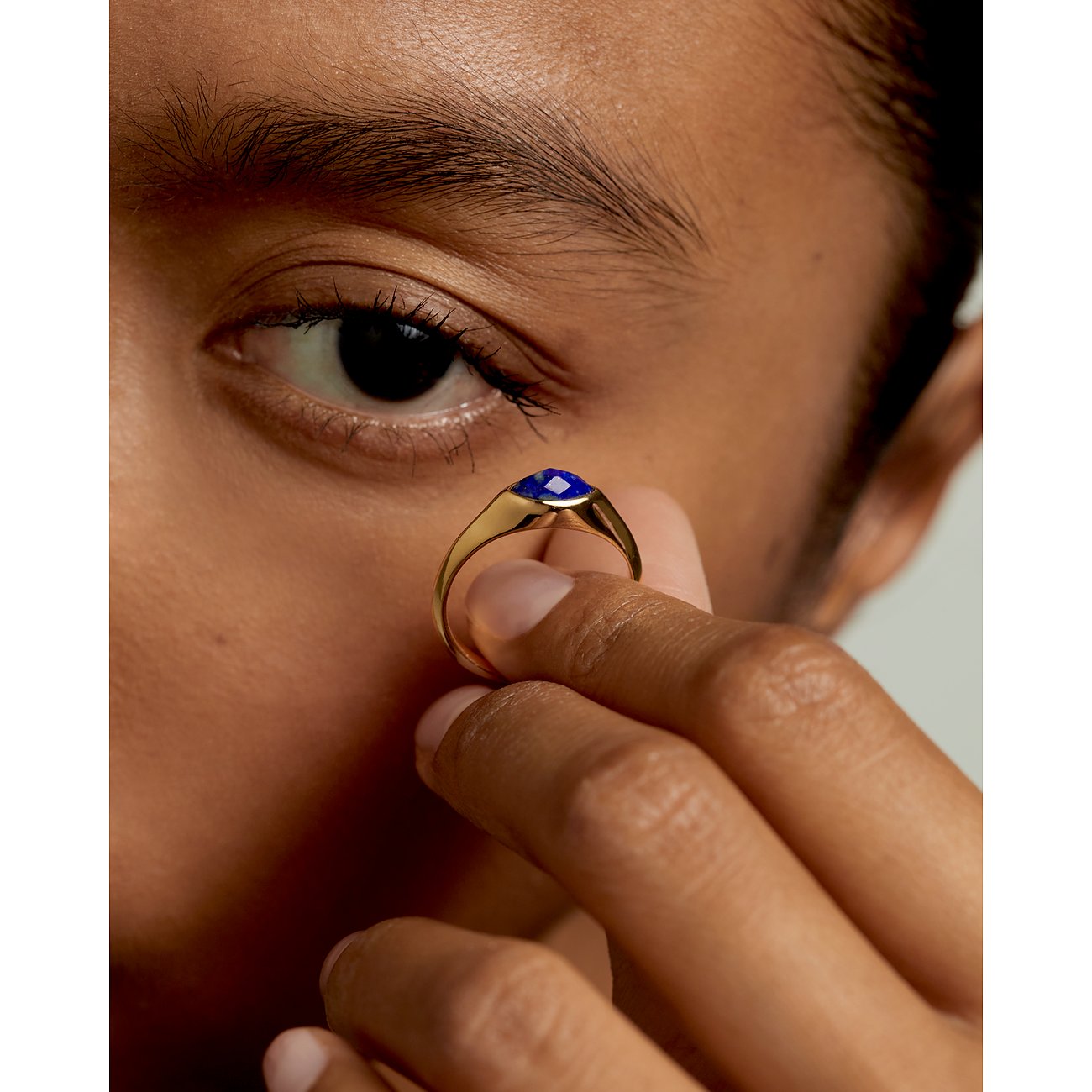 Nahaufnahme eines PdPaola Silberrings mit goldenem Finish und blauem Lapislazuli, gehalten vor einem Auge.
