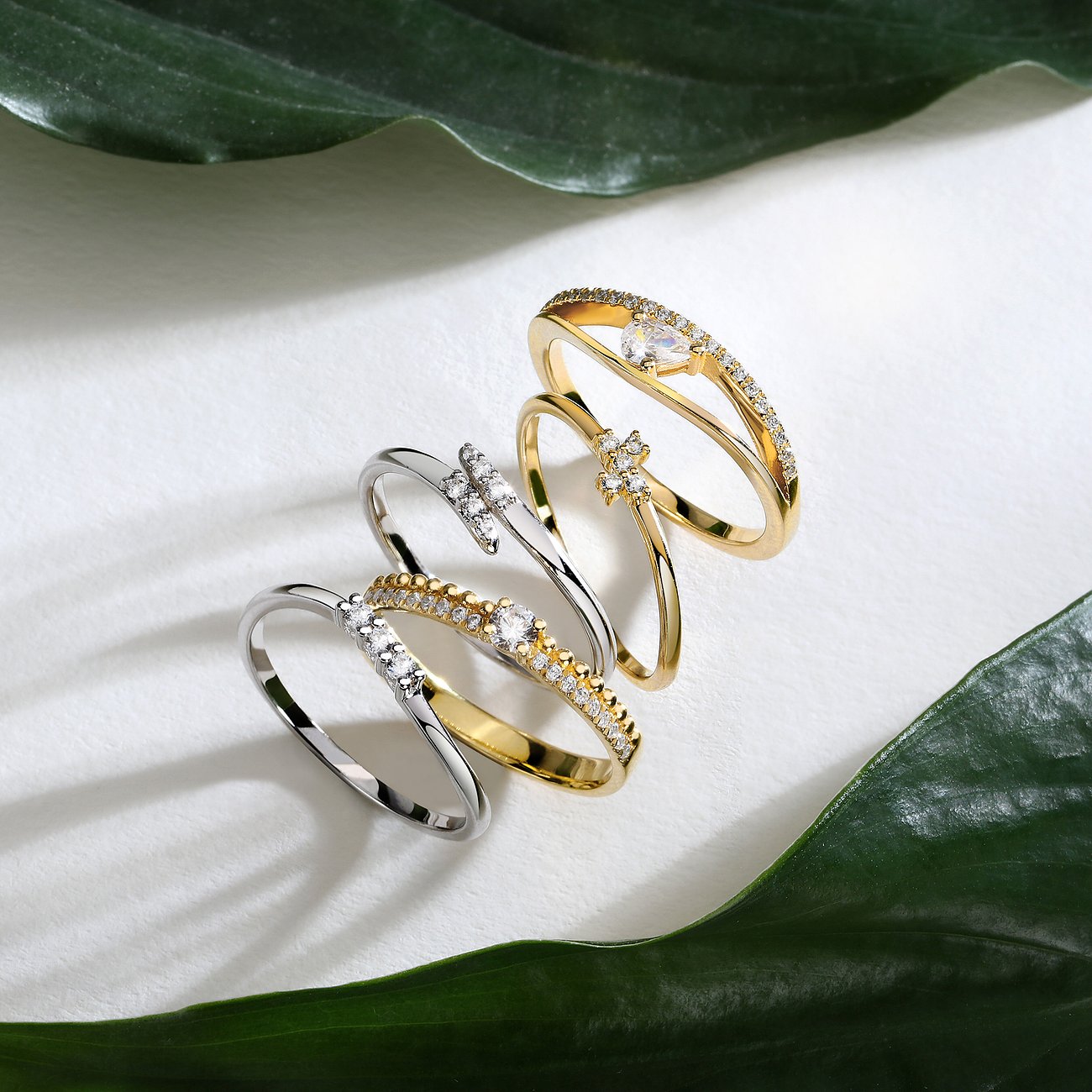 Live Diamond Ringe aus Gelbgold und silberfarbenem Metall, besetzt mit Diamanten, auf weißem Hintergrund mit grünen Blättern.