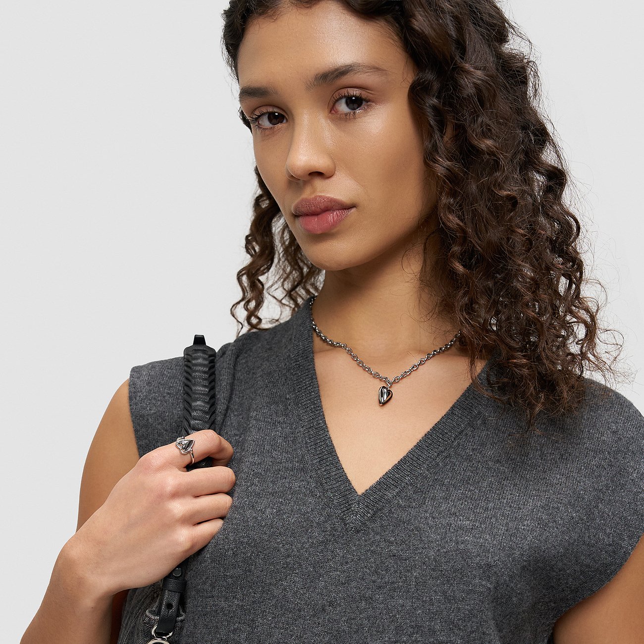Le modèle porte une bague Liebeskind en acier inoxydable et un collier cœur sur un haut gris.