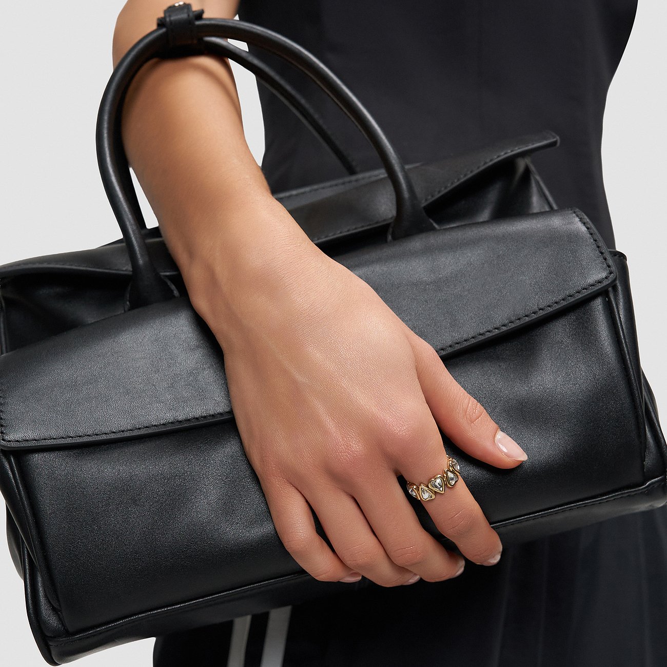 Nahaufnahme einer Hand, die eine schwarze Handtasche hält und einen Liebeskind Ring aus goldfarbenem Edelstahl trägt.