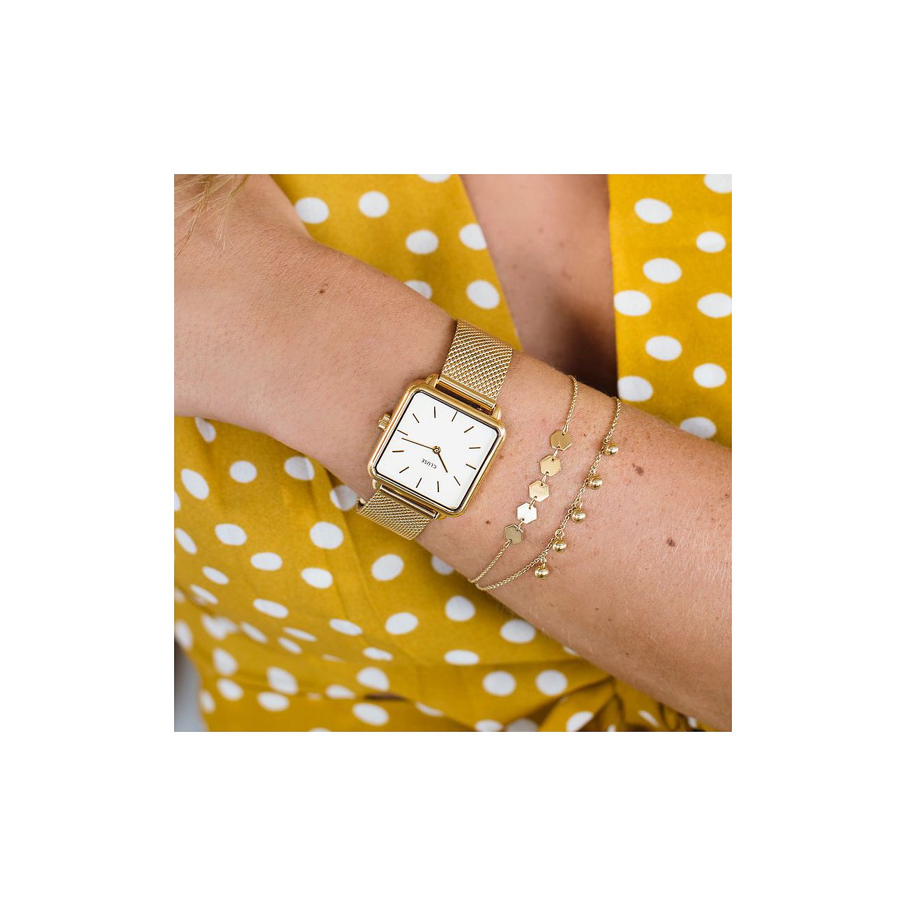 Am Handgelenk einer Frau ist eine Cluse Damenuhr aus goldfarbenem Metall mit quadratischem Zifferblatt zu sehen.