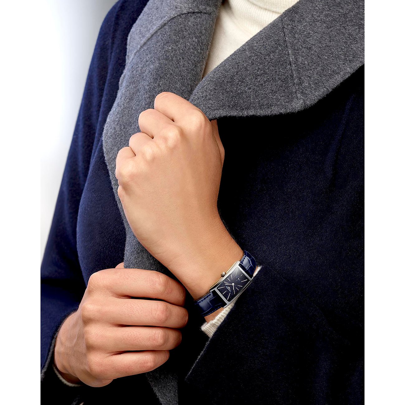 Eine Frau trägt eine rechteckige Longines Damenuhr aus Edelstahl mit marineblauem Lederarmband am Handgelenk.