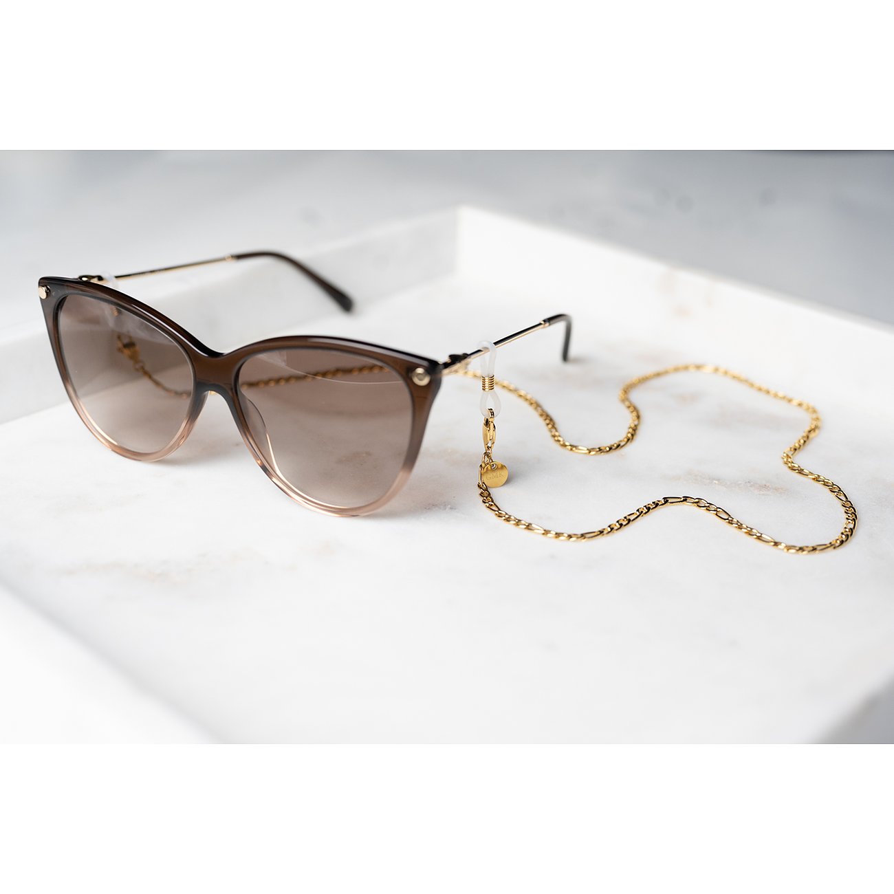 Eine Guido Maria Kretschmer Brillenkette aus goldfarbenem Edelstahl für die Sonnenbrille, auf einer weißen Marmorfläche.