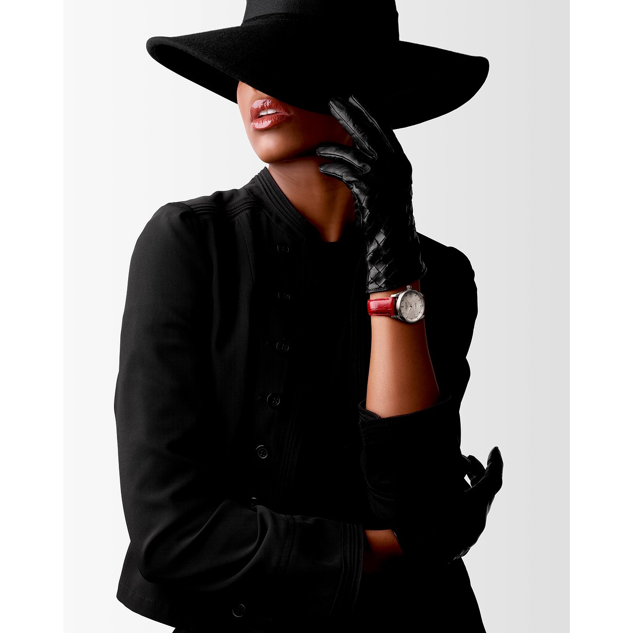 Eine Frau mit schwarzem Hut und Handschuhen trägt eine Longines Damenuhr aus Edelstahl mit Diamanten und roten Armband.