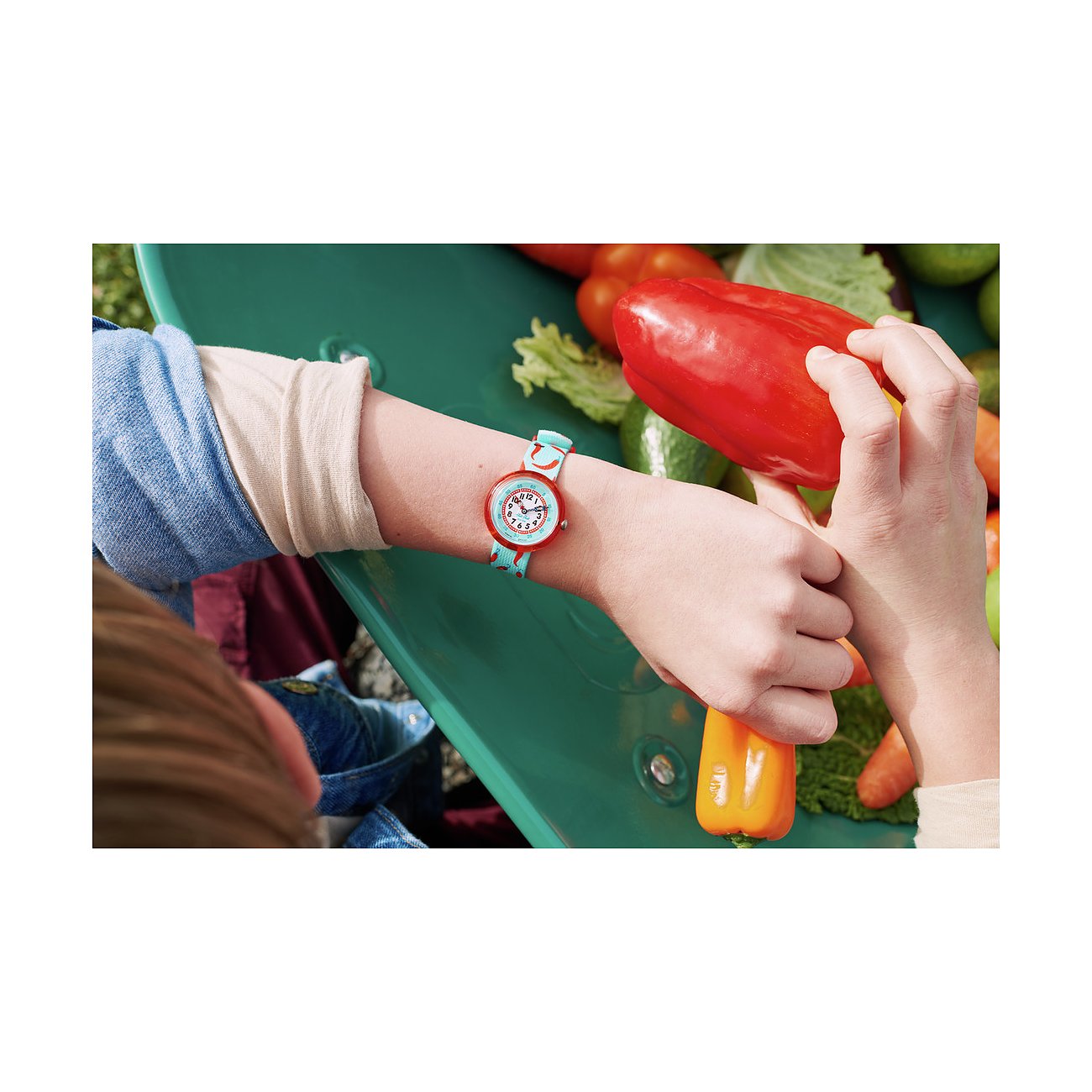 Montre enfant Flik Flak en plastique au poignet, cadran coloré, bracelet turquoise, jouant avec du poivron.