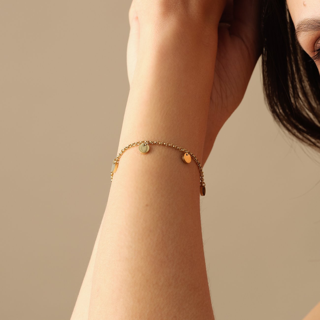 Ein glänzendes, goldenes FAVS Armband aus Edelstahl mit kleinen hängenden runden Scheiben, getragen an einem Arm.