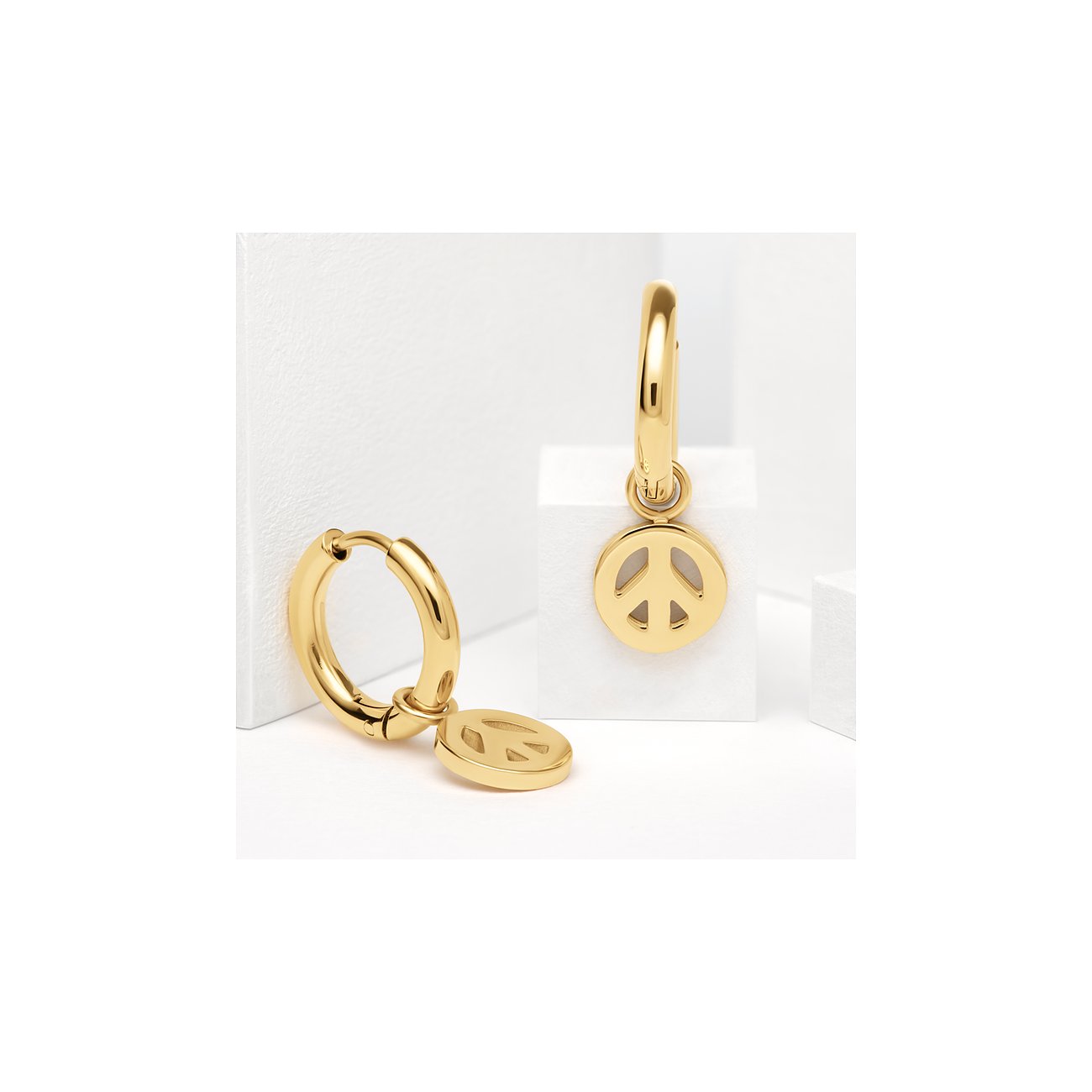 Guido Maria Kretschmer Ohrschmuck aus gelbvergoldetem Edelstahl mit Peace-Symbol-Anhänger, links liegend, rechts stehend.