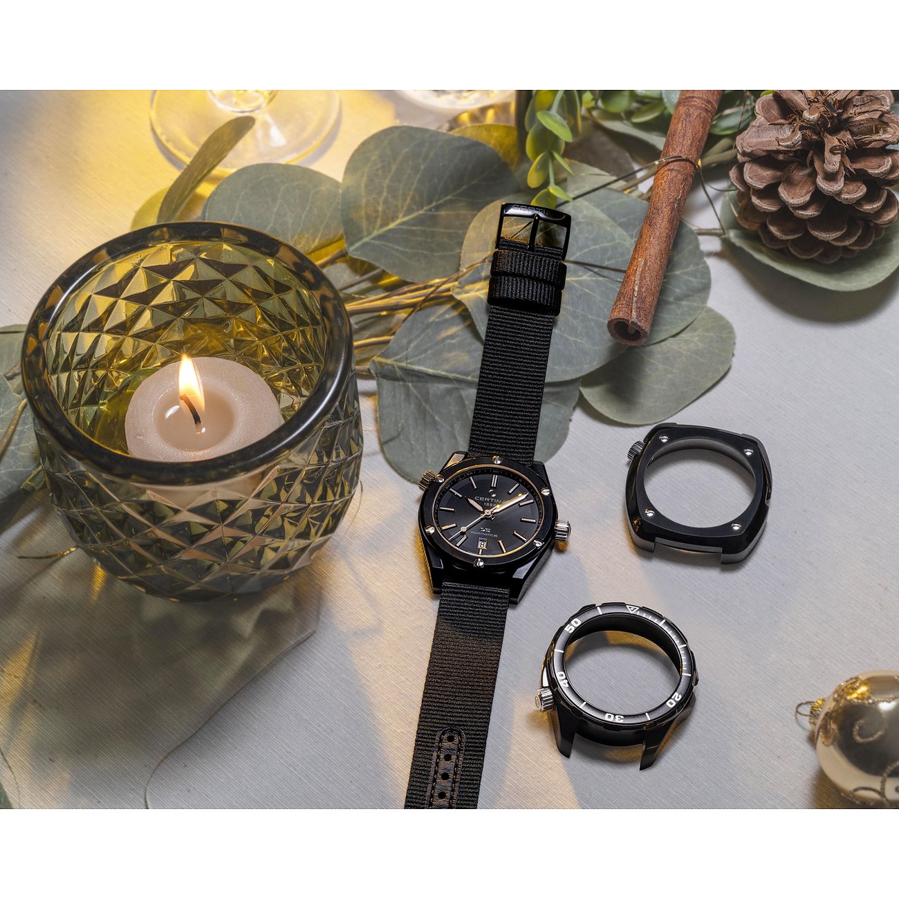 Certina Herrenuhr aus Edelstahl mit schwarzem Zifferblatt und Textilarmband, neben zwei Gehäusen und brennender Kerze auf Tuch.
