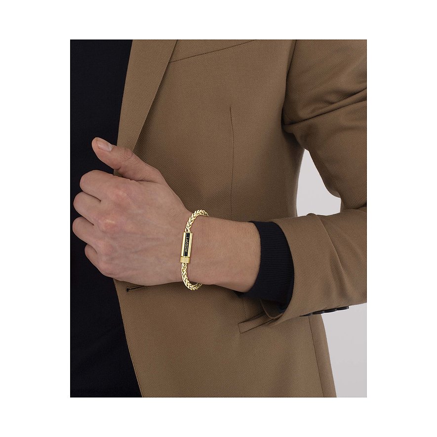 Een Lacoste armband van geelgoudkleurig roestvrij staal om de pols van een man met een bruine blazer.