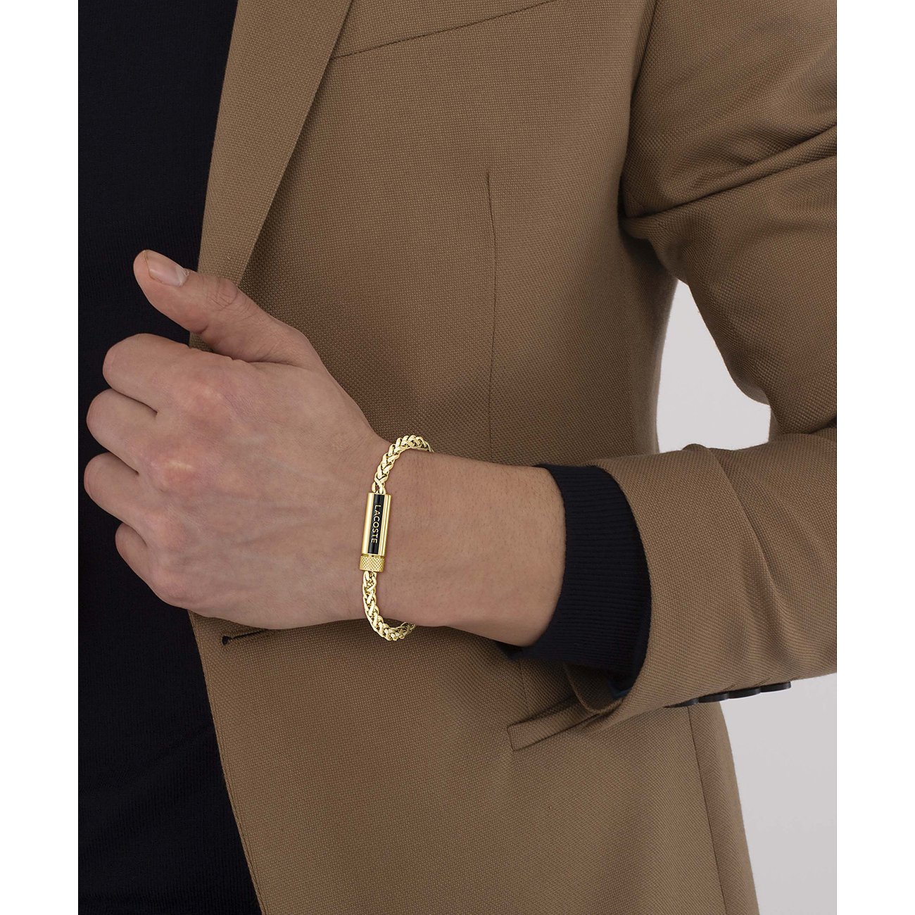 Een Lacoste armband van geelgoudkleurig roestvrij staal om de pols van een man met een bruine blazer.