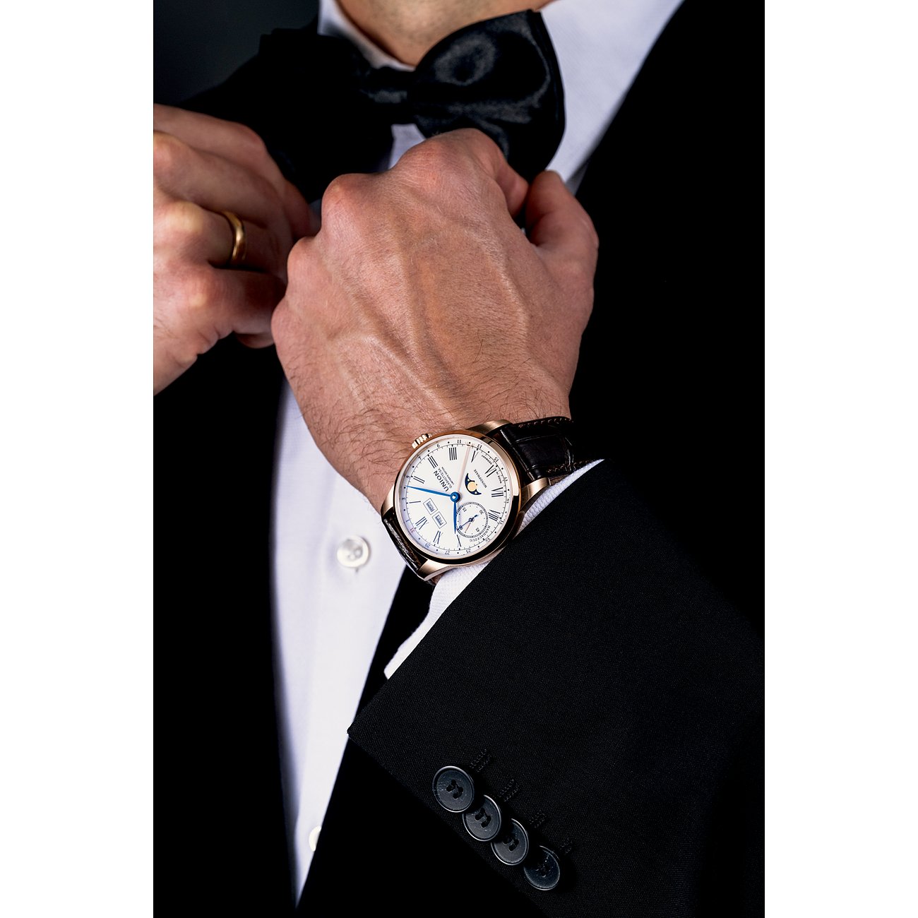 Ein Mann im Anzug bindet sich eine Fliege, am Handgelenk trägt er eine Union Glashütte Herrenuhr aus Roségold.