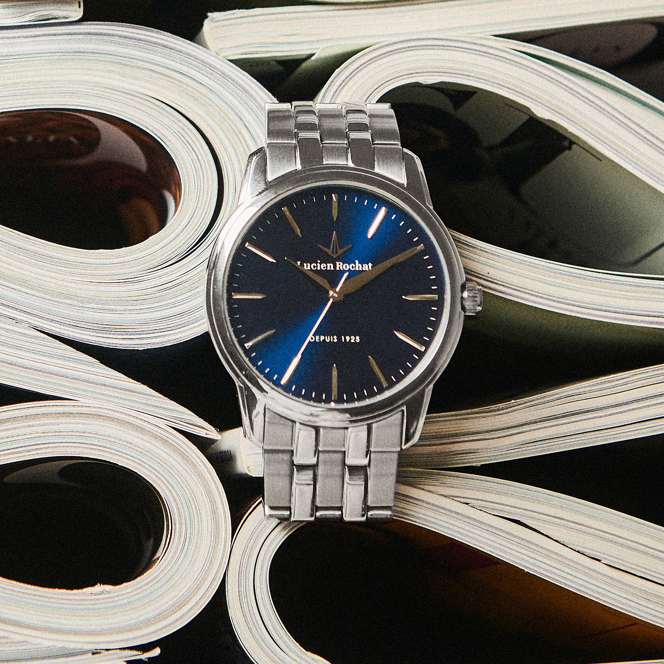 Une montre ronde Lucien Rochat pour homme en acier inoxydable avec cadran bleu, posée sur des magazines roulés.