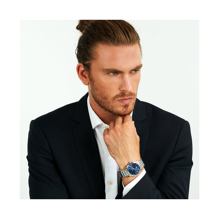 Un homme aux cheveux attachés porte une montre homme Lucien Rochat en acier inoxydable à cadran bleu.