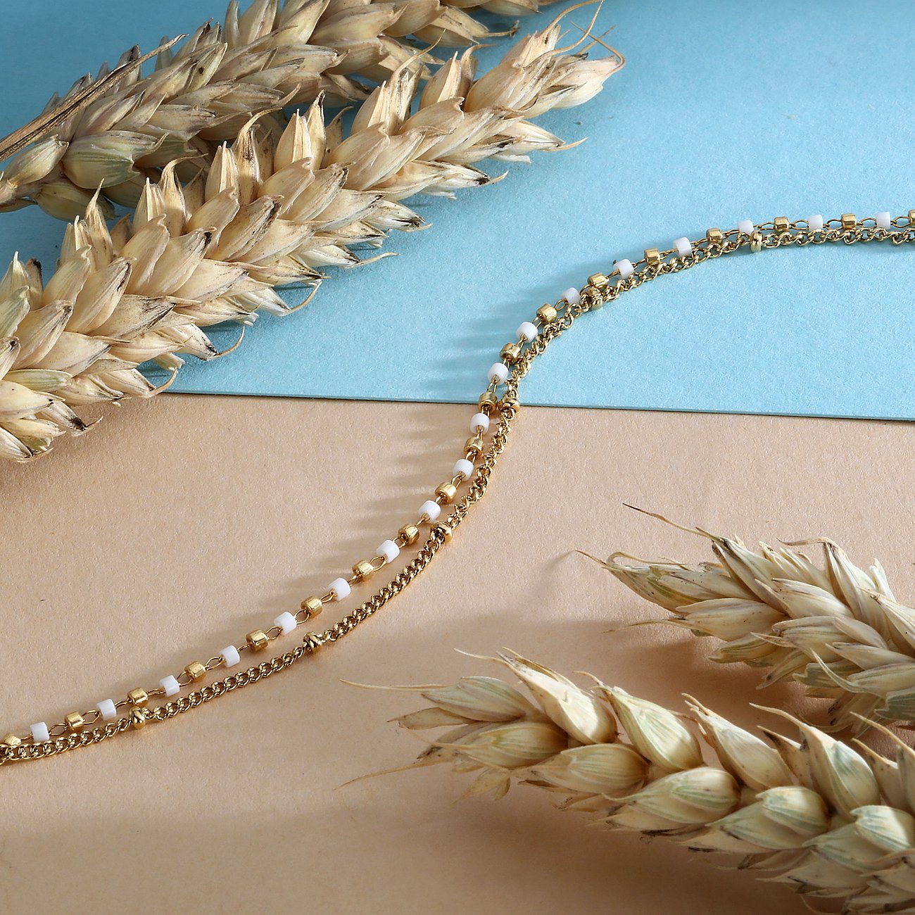 La Petite Story Armschmuck aus gelb-goldenem Edelstahl und weißer Emaille auf beigem und blauem Hintergrund.