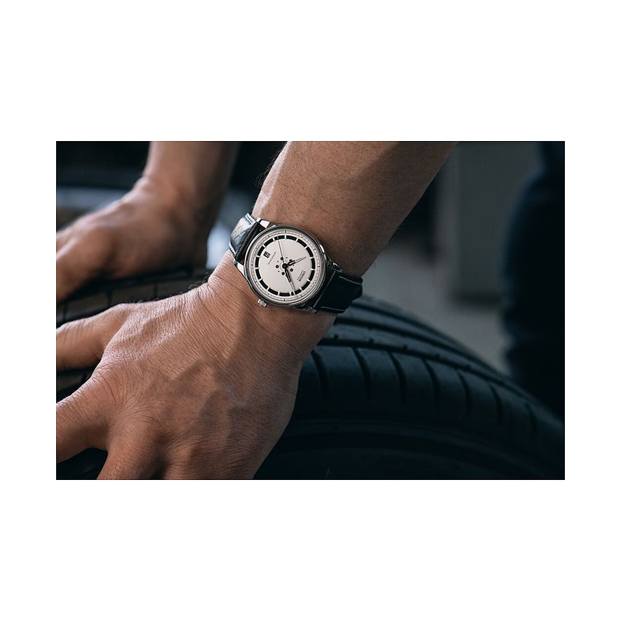 Am Handgelenk eines Mannes, dessen Hände auf Autoreifen ruhen, sitzt eine Union Glashütte Herrenuhr aus Edelstahl.