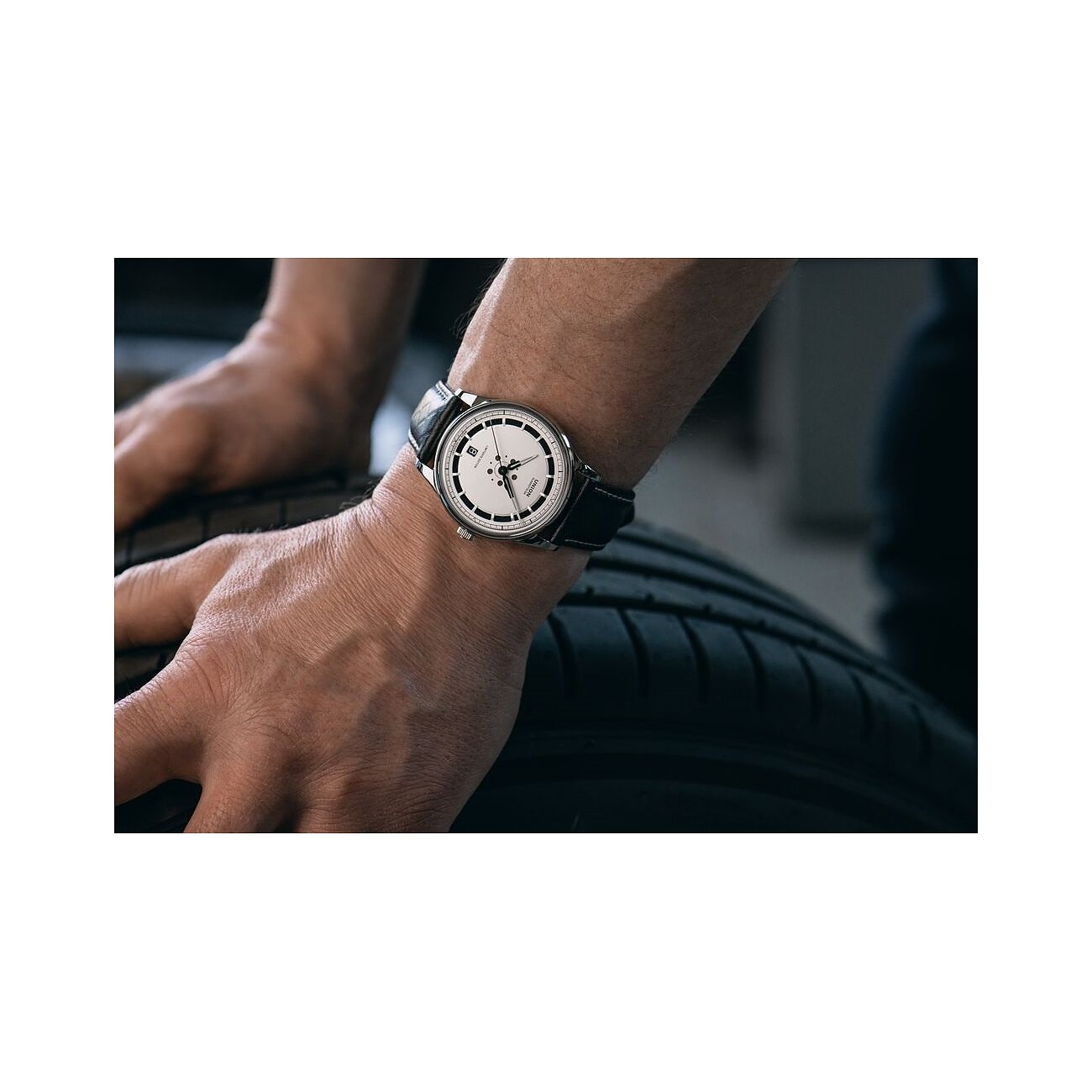 Am Handgelenk eines Mannes, dessen Hände auf Autoreifen ruhen, sitzt eine Union Glashütte Herrenuhr aus Edelstahl.