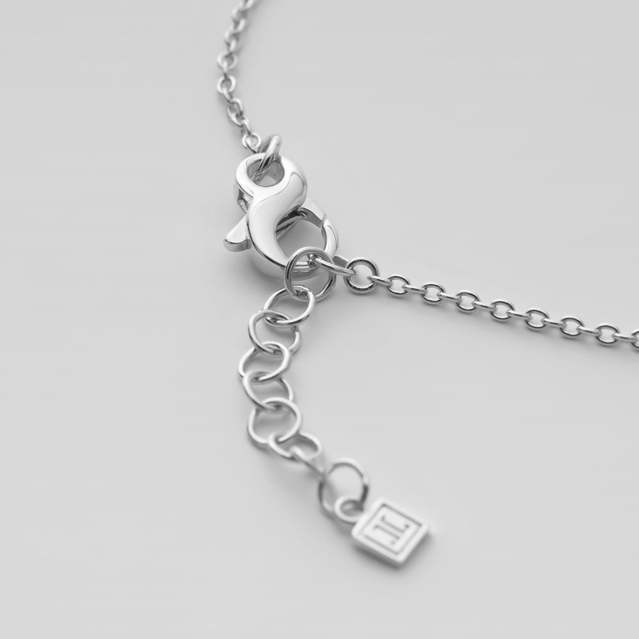 Nærbillede af lukkemekanismen og forlængerkæden på et sølvfarvet JETTE armbånd med et lille mærkevedhæng.