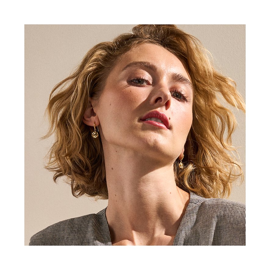 Modèle avec boucles d'oreilles pendantes Skagen dorées en acier inoxydable, brillantes en gros plan, encadrant le visage.
