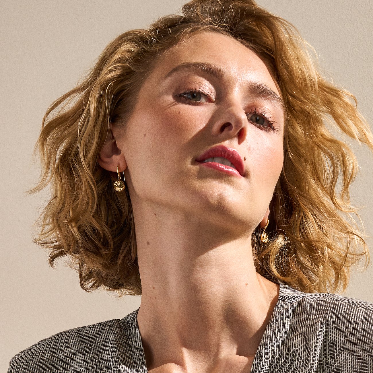 Modèle avec boucles d'oreilles pendantes Skagen dorées en acier inoxydable, brillantes en gros plan, encadrant le visage.