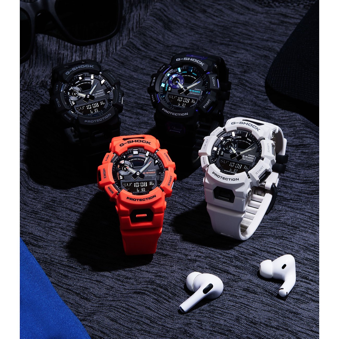 Eine Nahaufnahme zeigt vier Casio Herrenuhren aus Resin in Schwarz, Lila, Orange und Weiß mit Airpods auf dunklem Stoff.