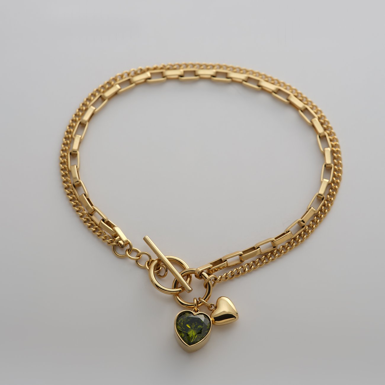 Guido Maria Kretschmer Armband aus goldfarbenem Edelstahl. Es hat zwei Kettenstränge und einen grünen Zirkonia-Anhänger.