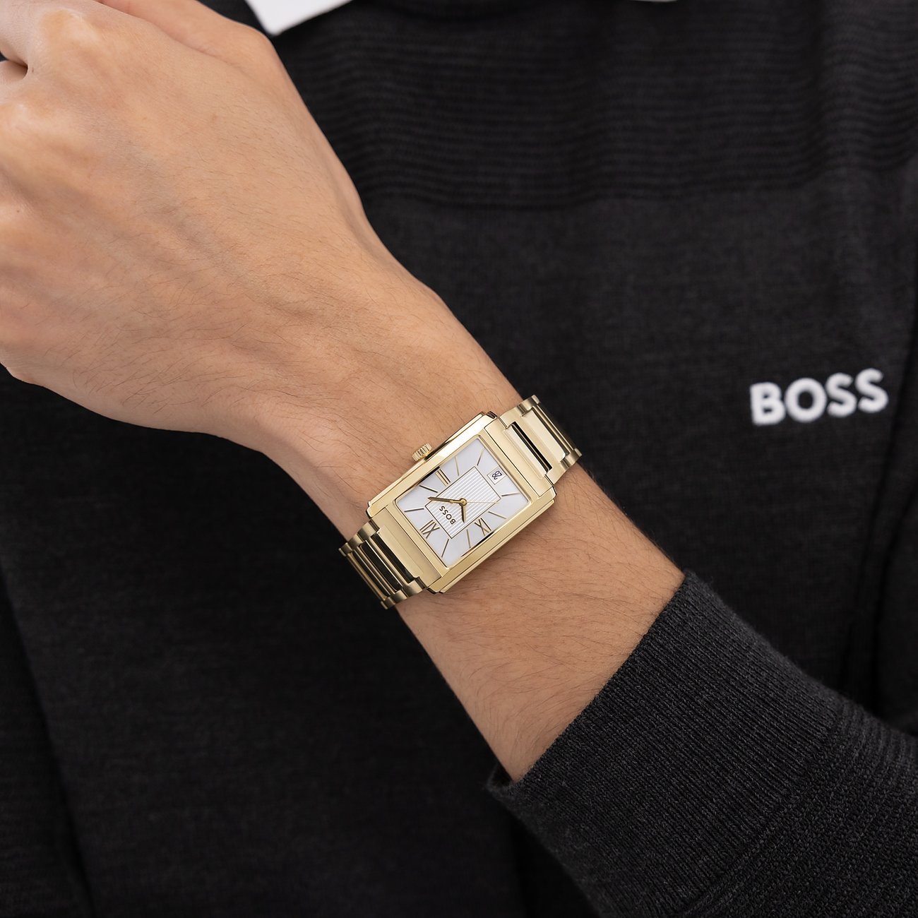 Am Handgelenk eines Mannes wird eine goldfarbene Boss Herrenuhr aus Edelstahl mit rechteckigem Zifferblatt gezeigt.