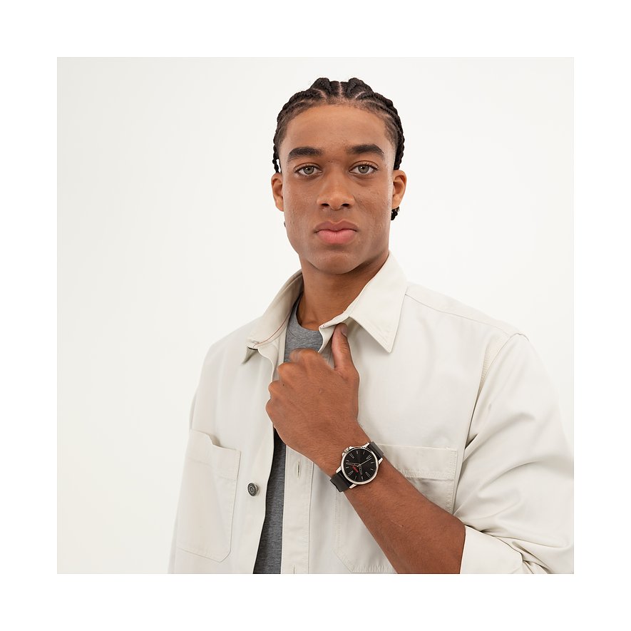 Ein Mann mit geflochtenen Haaren und einem beigen Hemd posiert mit dem Hugo Herrenuhren-Modell aus Edelstahl am Handgelenk.