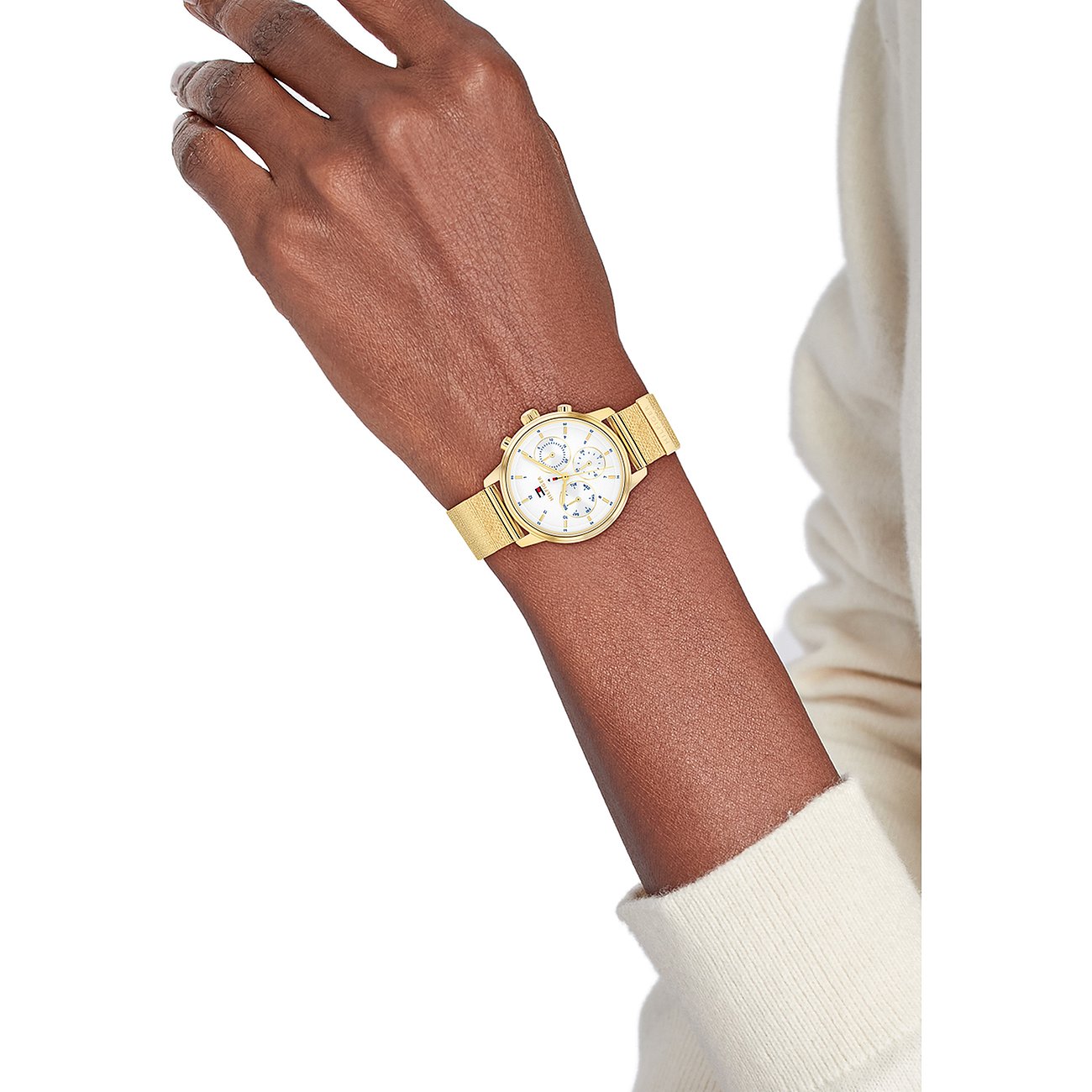 Tommy Hilfiger Damenuhr aus goldfarbenem Edelstahl mit weißem Zifferblatt und Mesh-Armband am Handgelenk getragen.