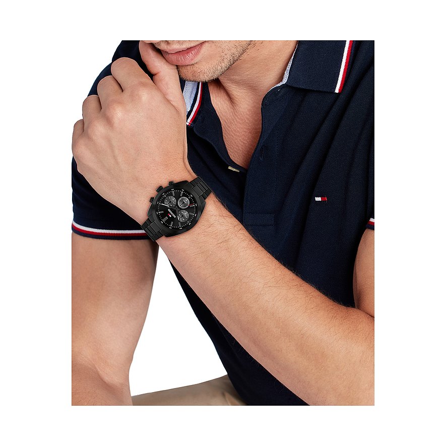 Een man in een poloshirt draagt het Tommy Hilfiger herenhorloge van zwart roestvrij staal om zijn pols.