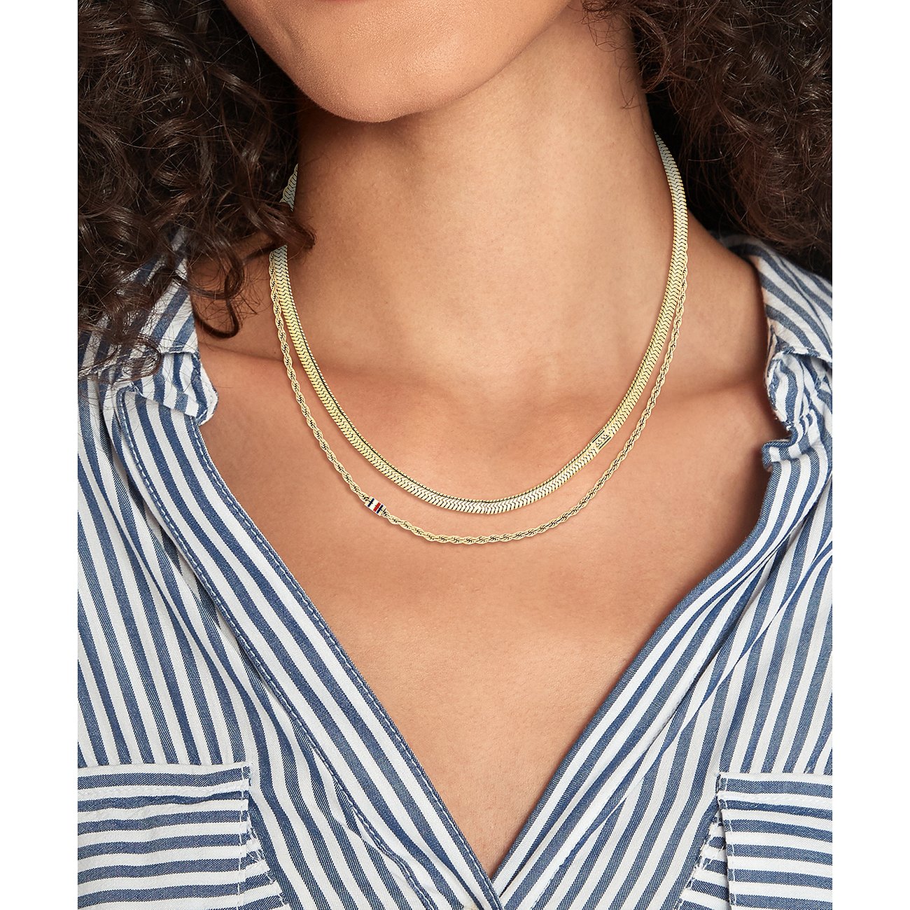 Tommy Hilfiger Halsschmuck aus goldfarbenem Edelstahl. Eine Frau trägt eine mehrreihige Kette auf ihrem gestreiften Hemd.