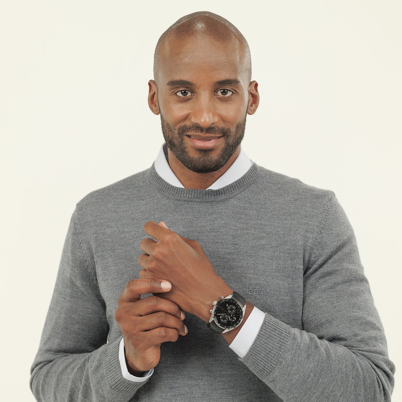 Lächelnder Mann mit Boss Herrenuhr aus Edelstahl am linken Handgelenk, grauem Pullover und weißem Hemd.