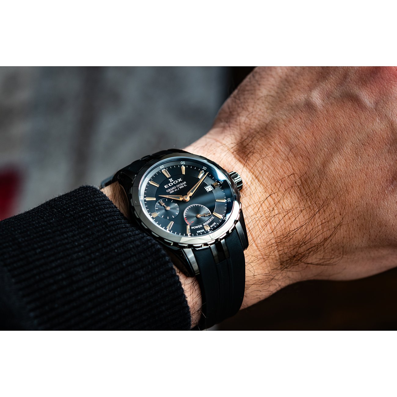 Die Edox Herrenuhr aus Edelstahl und Aluminium mit schwarzem Zifferblatt und Kautschukarmband wird am Handgelenk getragen.
