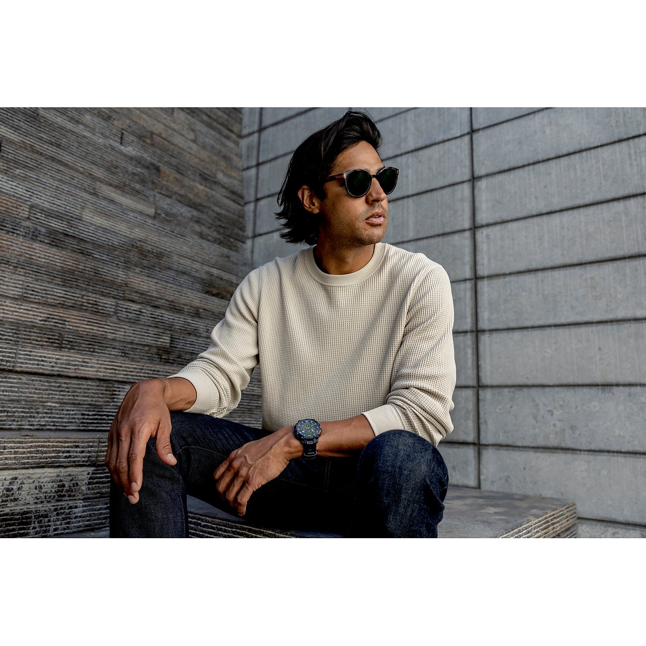 Ein Mann trägt eine Garmin Smartwatch aus Kunststoff und sitzt mit Sonnenbrille und Jeans lässig auf einer Treppe.