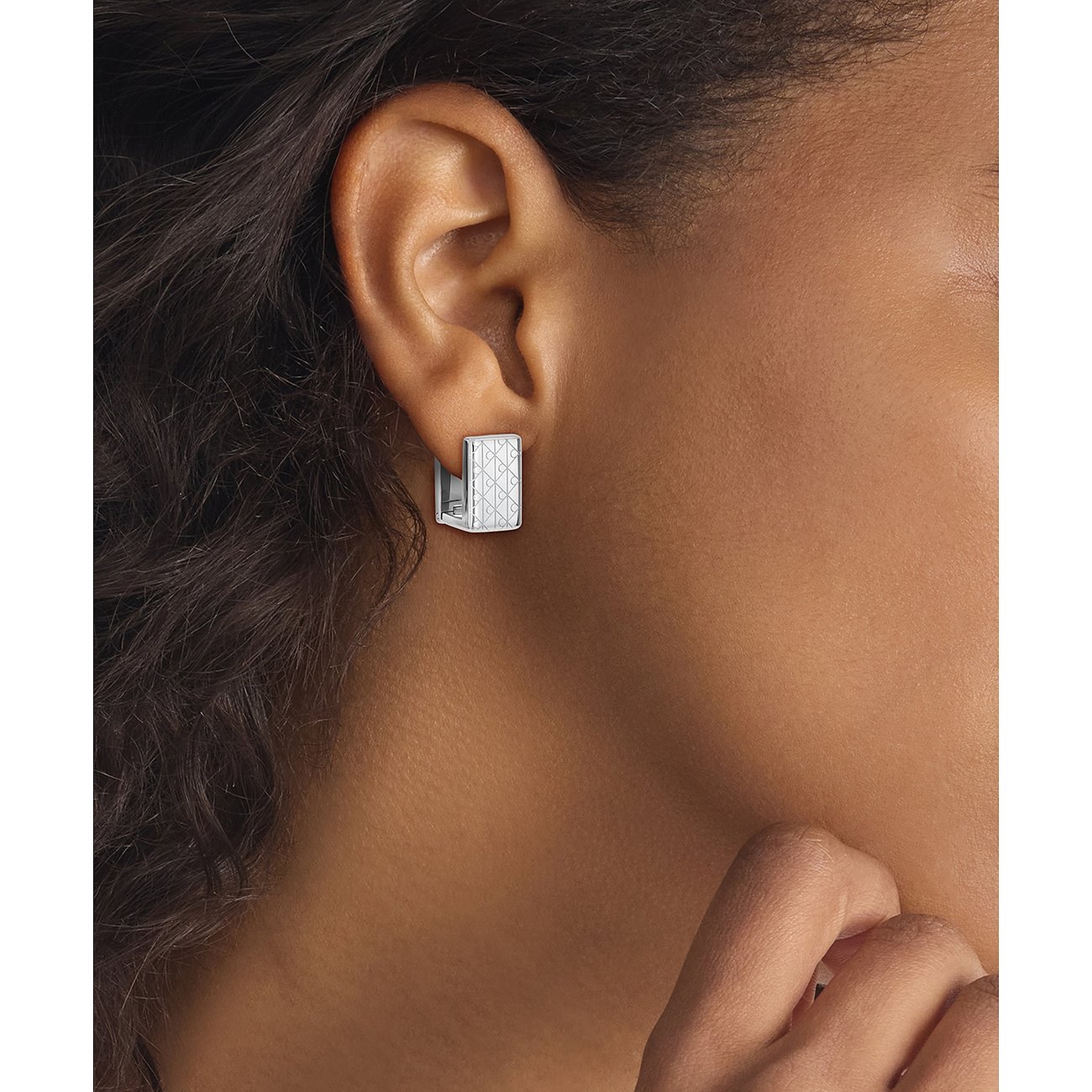 Ein Calvin Klein Ohrring aus Edelstahl ist am Ohr getragen zu sehen. Er ist halbmondförmig mit einem Muster.