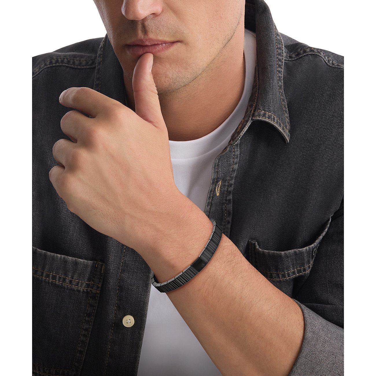 Nahaufnahme eines Mannes, der ein Calvin Klein Armband aus Edelstahl trägt, Arm auf dem Schoß, Hand am Kinn.