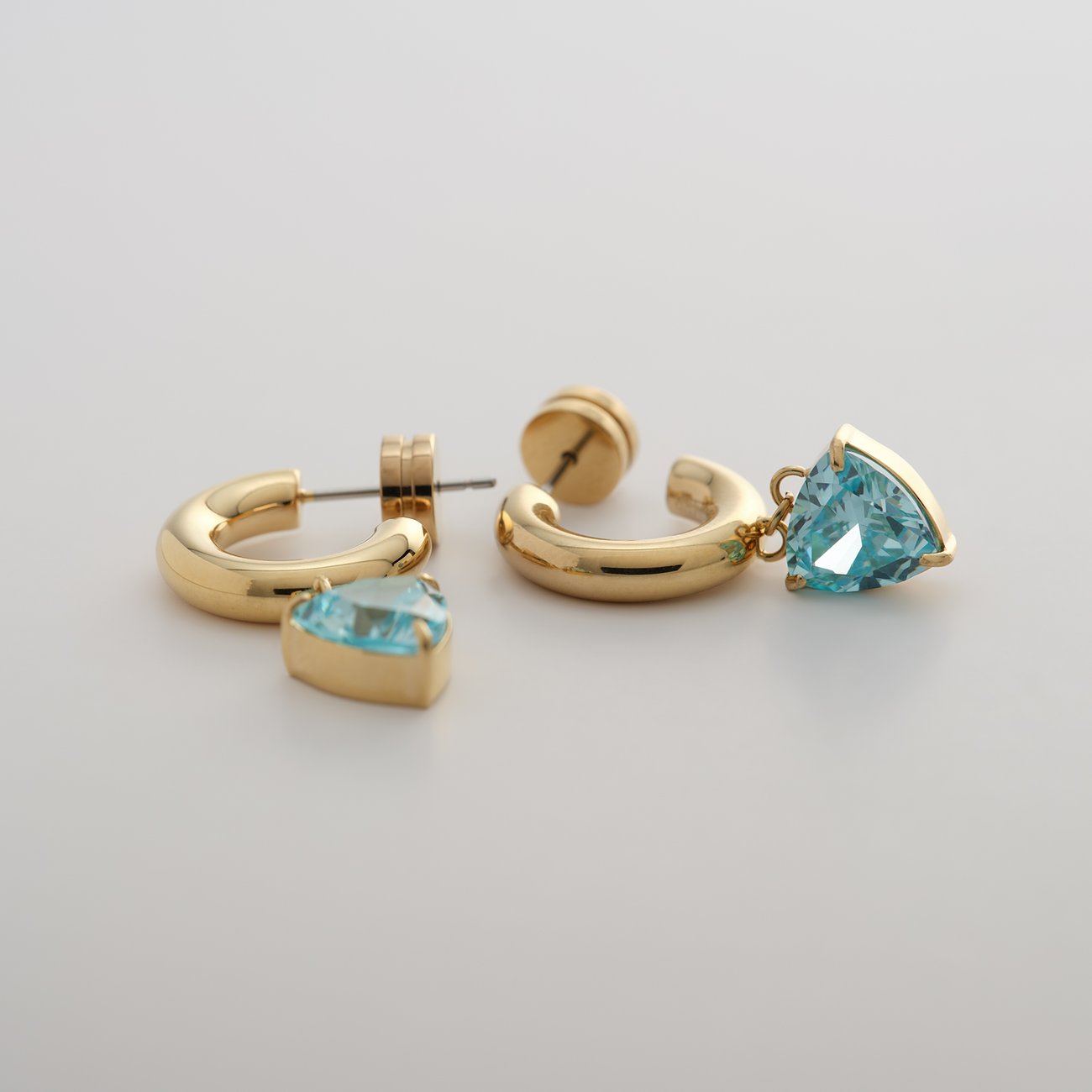 Zwei Guido Maria Kretschmer Ohrringe aus goldfarbenem Edelstahl mit hellblauen Zirkonia-Farbsteinen.