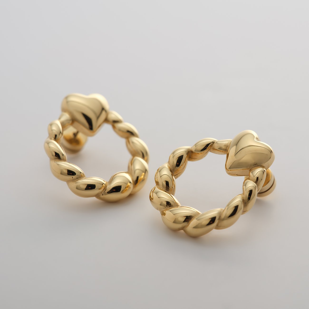 Zwei Guido Maria Kretschmer Ohrringe aus goldfarbenem Edelstahl, die wie gedrehte Creolen mit Herzdetails aussehen.