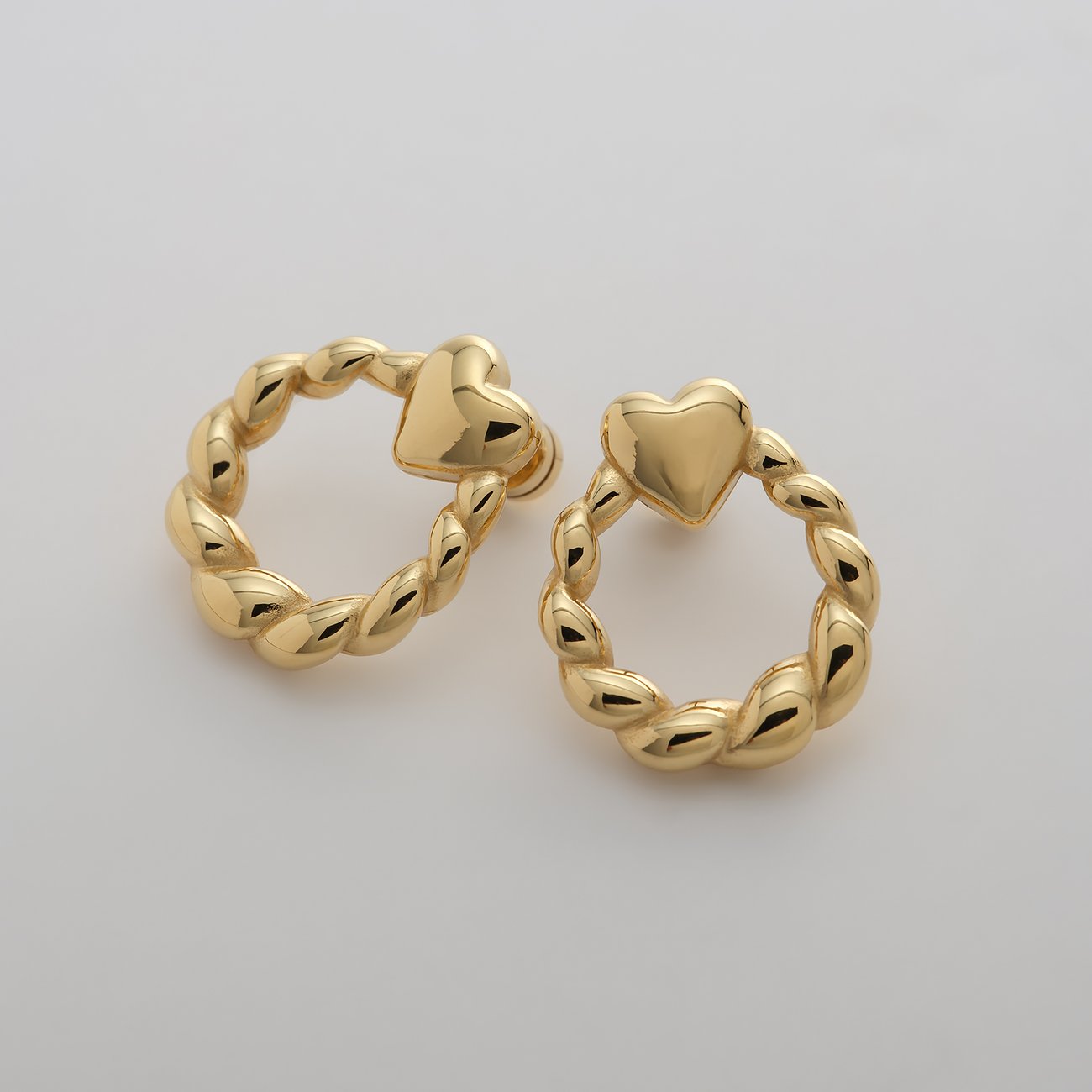 Zwei Guido Maria Kretschmer Herz Ohrringe aus goldenem Edelstahl gefertigt, auf einem hellen Hintergrund.