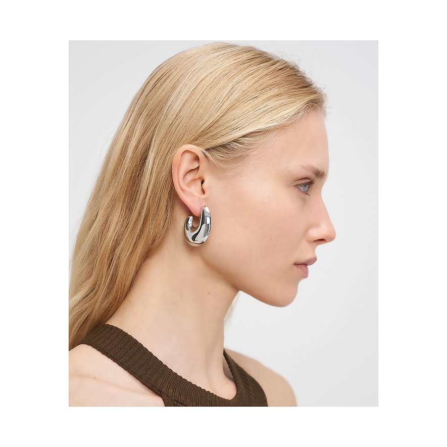 Close-up van een zilverkleurig Liebeskind roestvrijstalen oorsieraad aan een vrouwenoor, zijwaarts gefotografeerd.