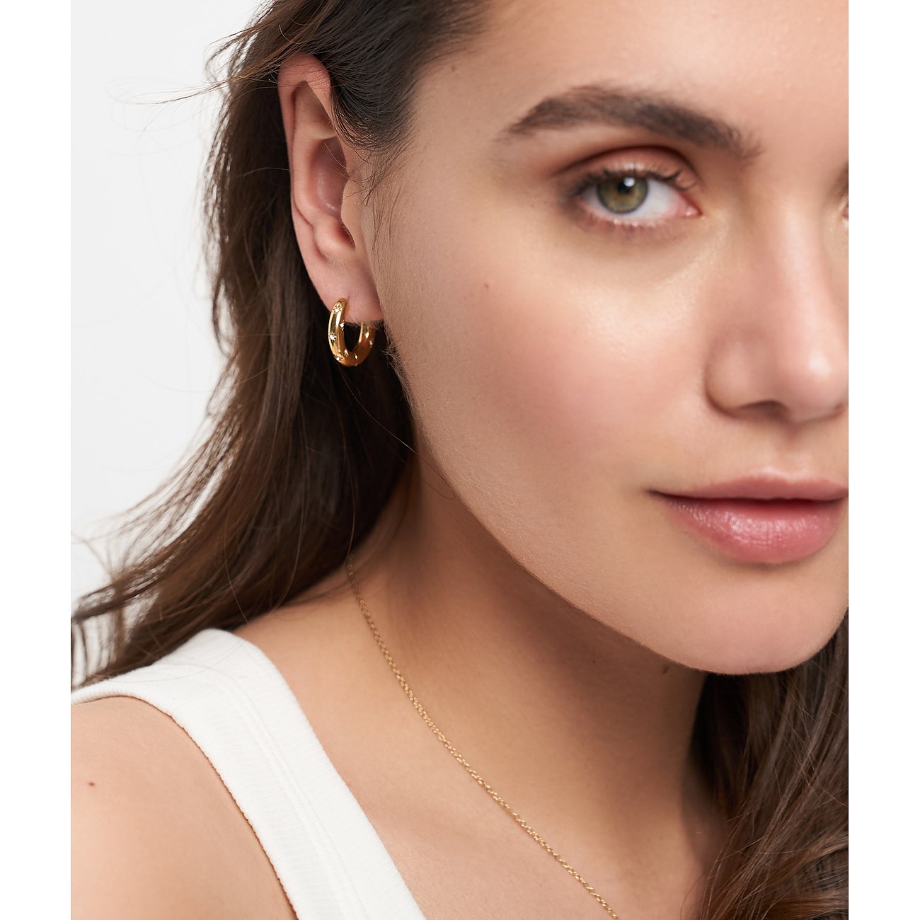 Närbild: Kvinna med allvarlig blick, bär Tamaris örhänge i guldfärgat rostfritt stål, smyckat med gnistrande glasstenar.