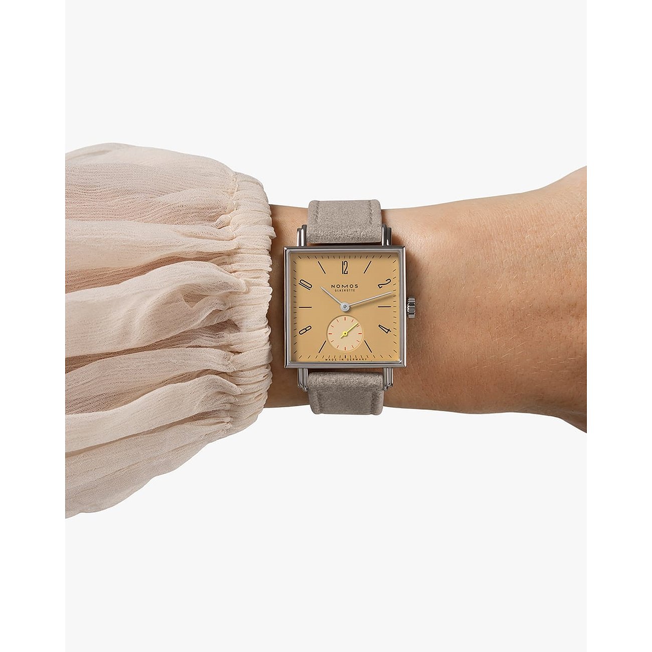 Nomos Glashütte Damenuhr aus Edelstahl mit eckigem, beigem Zifferblatt und grauem Band am Handgelenk, Nahaufnahme.