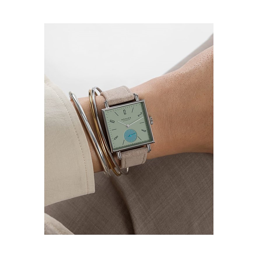 Am Handgelenk getragene Nomos Glashütte Damenuhr aus Edelstahl mit hellem Zifferblatt und beigem Band.