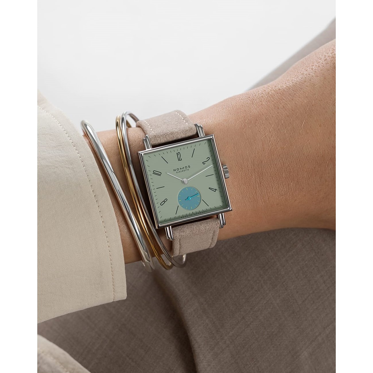 Am Handgelenk getragene Nomos Glashütte Damenuhr aus Edelstahl mit hellem Zifferblatt und beigem Band.