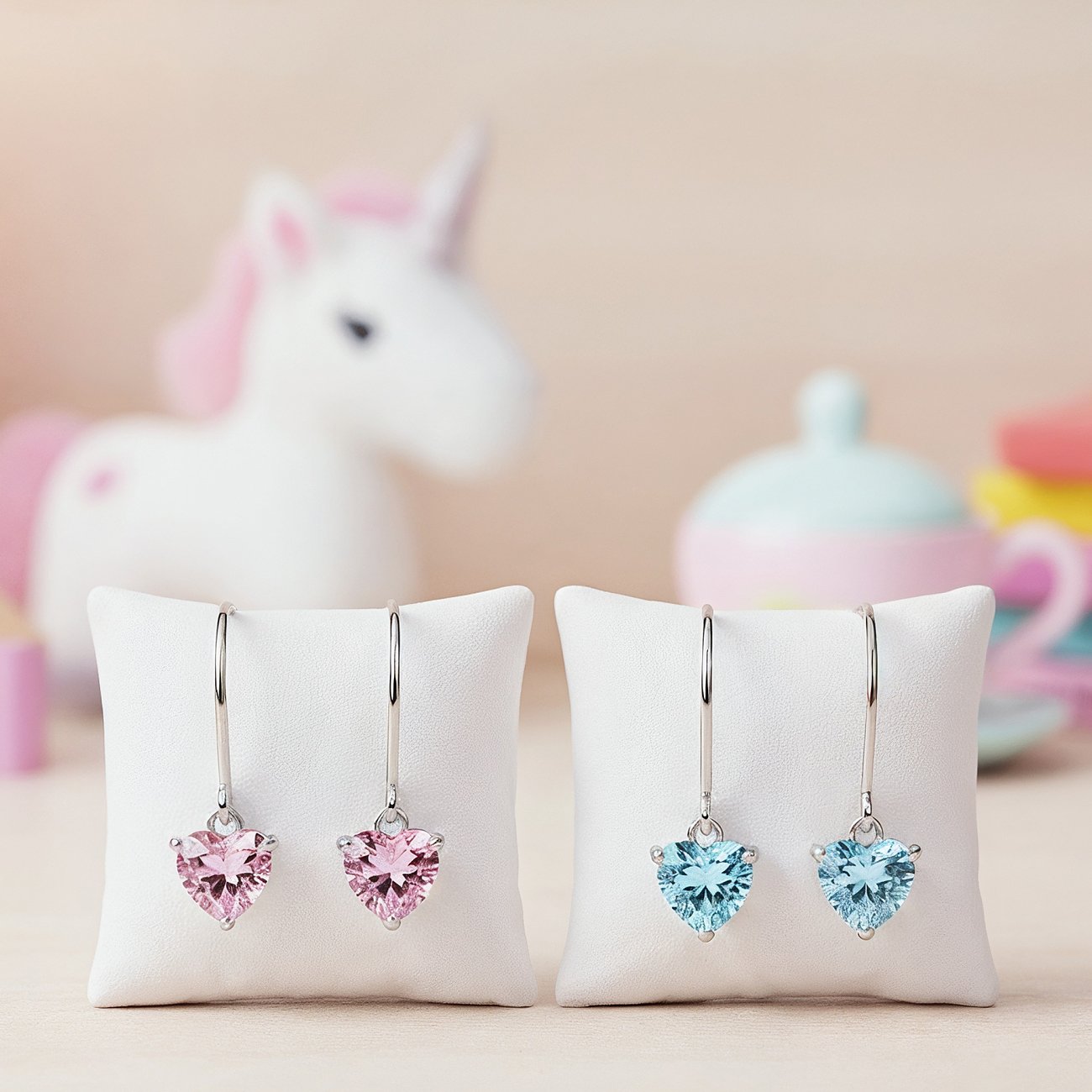Engelsrufer Ohrhänger aus Edelstahl mit herzförmigen Zirkonia-Farbsteinen in Rosa und Hellblau auf weißen Kissen.
