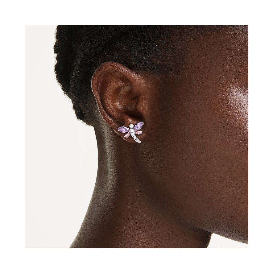 Een Swarovski libelle-oorknopje van metaal met violette en roze edelstenen, gedragen aan één oor.