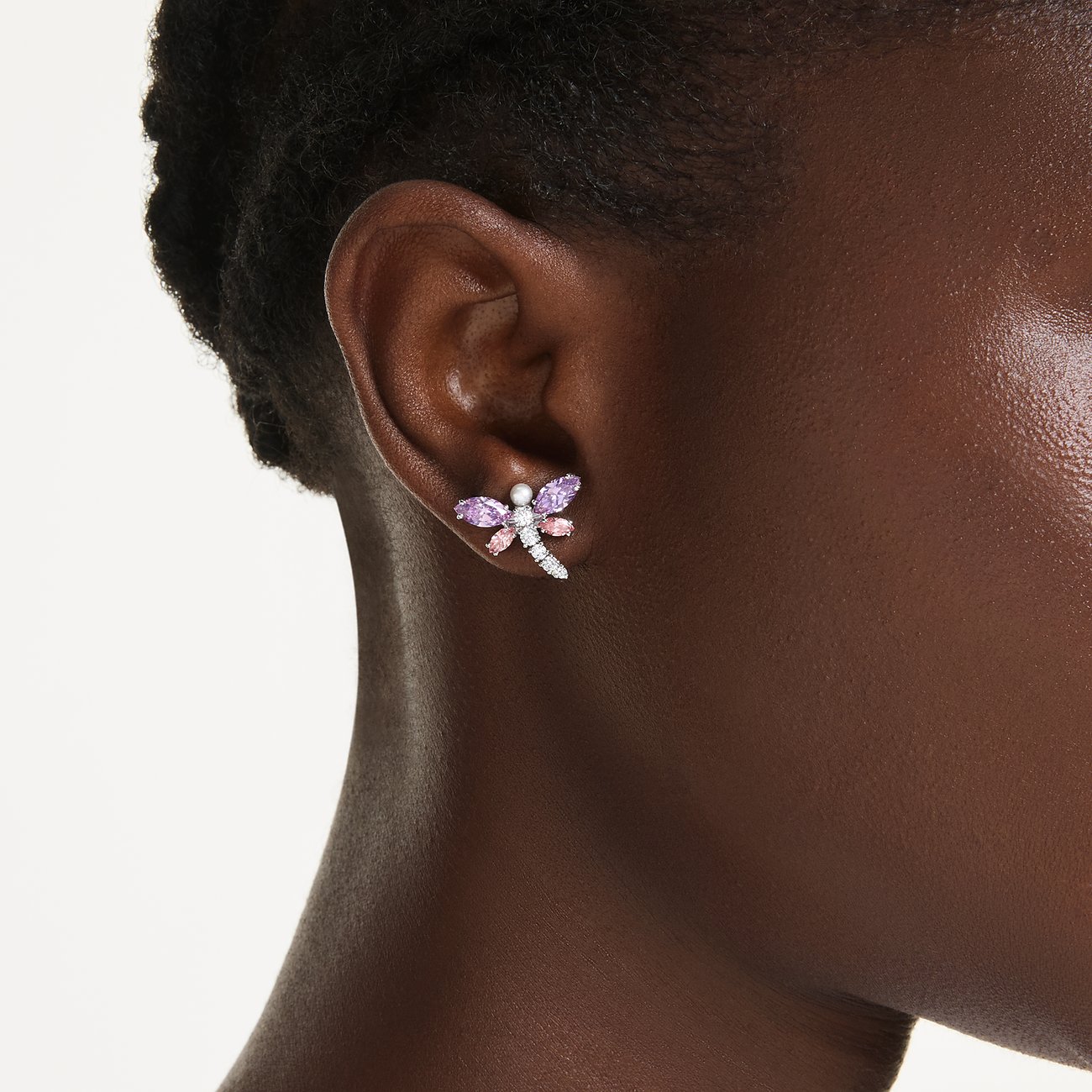 Een Swarovski libelle-oorknopje van metaal met violette en roze edelstenen, gedragen aan één oor.