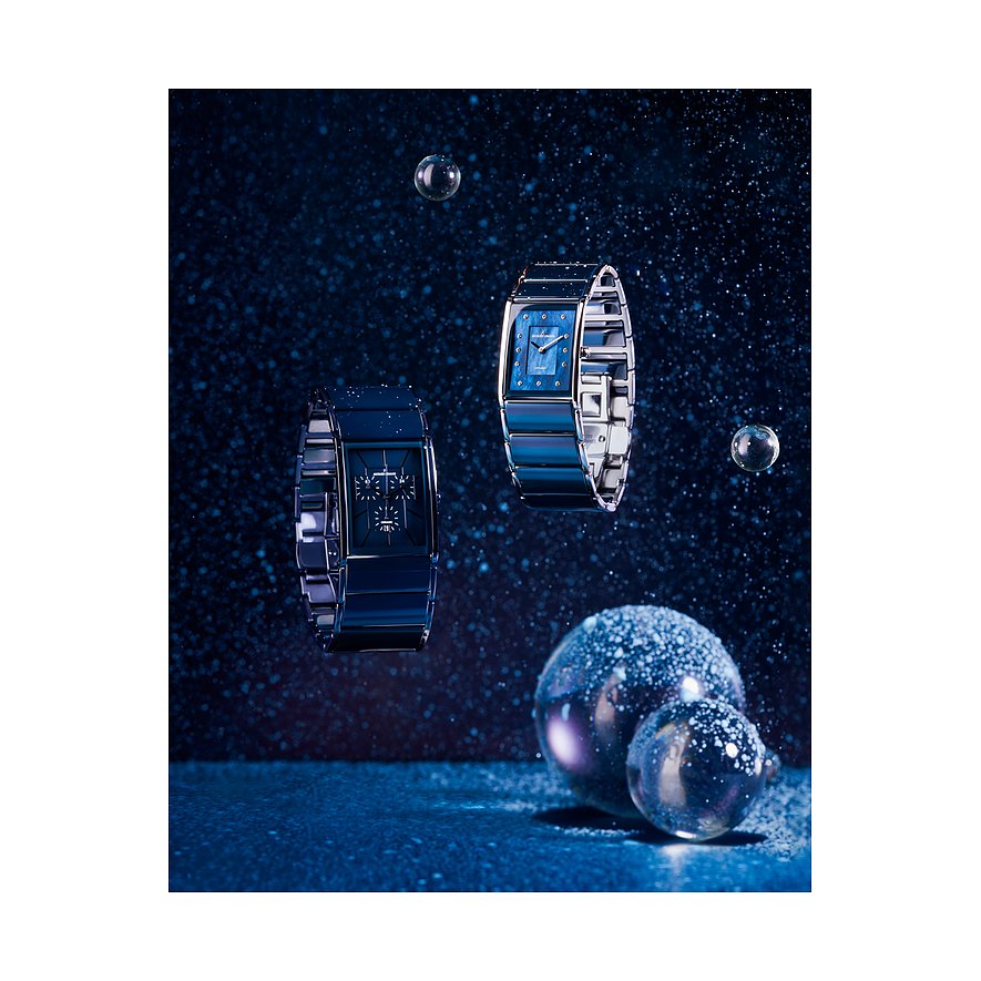 Zwei schwebende Jacques Lemans Damenuhren aus blau gefärbtem Edelstahl vor dunklem Hintergrund mit glitzernden Details.