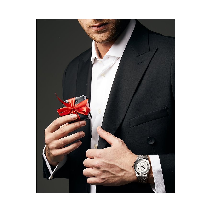 Ein Mann im schwarzen Anzug mit einer Jacques Lemans Herrenuhr aus Edelstahl am Handgelenk hält ein kleines Geschenk.