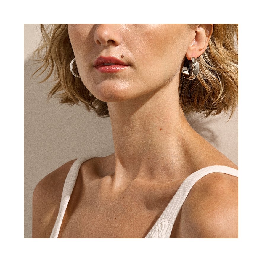 Une femme porte des boucles d'oreilles Skagen en acier inoxydable. Le mannequin blond a les cheveux courts et un haut blanc.