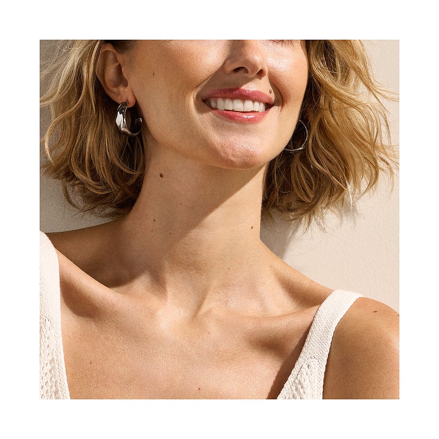Gros plan d'une femme souriante portant une paire de boucles d'oreilles créoles Skagen brillantes en acier inoxydable.